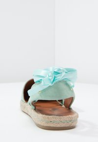 Sandalia de espadrille de ante verde menta con suela de yute trenzado. Presenta un lazo decorativo de cinta azul claro en el tobillo y acentos en tono dorado.
