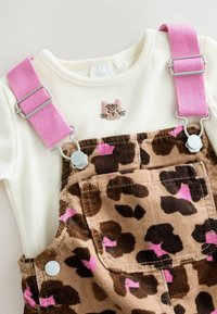 Cremig-rippiges Langarmshirt mit gesticktem Katzenkopf, kombiniert mit khaki- und braunen, mit Blumenmuster versehenen Cordoveralls mit verstellbaren rosa Trägern.