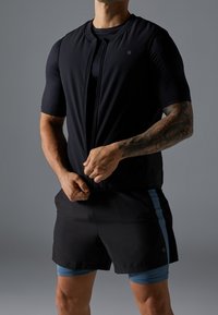 Man med tatuerad arm som bär svarta träningsshorts och tröja, drar upp en svart ärmlös väst mot en grå bakgrund.