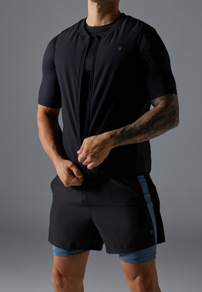 Man med tatuerad arm som bär svarta träningsshorts och tröja, drar upp en svart ärmlös väst mot en grå bakgrund.