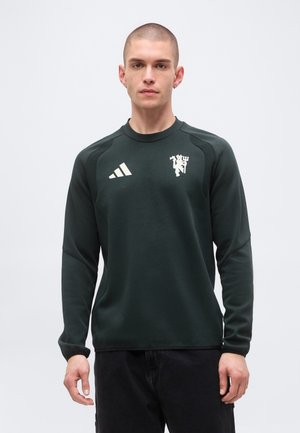Jeune homme aux cheveux courts portant un maillot de sport foncé à manches longues avec des logos blancs sur la poitrine, debout devant un fond uni.