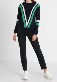 Granatowy sweter z zielono-białym wzorem w kształcie litery V, o prążkowanej fakturze, z długimi rękawami, zestawiony z czarnymi spodniami i białymi tenisówkami.