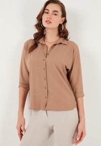 Beige button-up shirt with a textured fabric, a collar, and rolled 3/4 sleeves, paired with light-colored pants.