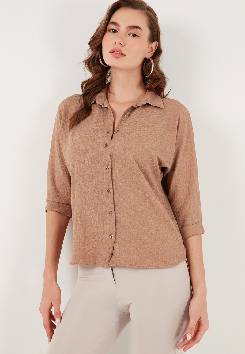 Beige button-up shirt with a textured fabric, a collar, and rolled 3/4 sleeves, paired with light-colored pants.