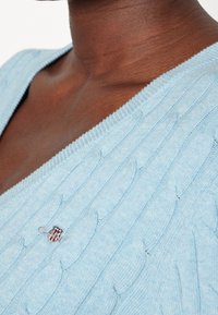 Zbliżenie ciemnoskórej osoby w jasnoniebieskim swetrze w warkoczowym splocie z małym haftowanym logo w pobliżu dekoltu.