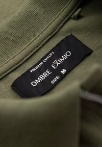 Olivi vihreä kangaskaulus, jossa on musta etiketti, johon on kirjattu "OMBRE EXIMIO", mikä osoittaa premium-laatua ja keskikokoista, sileäpintaista materiaalia.