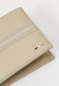 Beige skinnlommebok med teksturerte bånd, sydd kanter, og preget "BOSS"-logo. Har fargeaksenter i svart, hvitt og kobber.