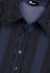 Chemise bleu foncé avec bordure en dentelle noire le long de la patte de boutonnage et du col, avec quatre boutons et une étiquette "THE KOOPLES" à l'intérieur du col.