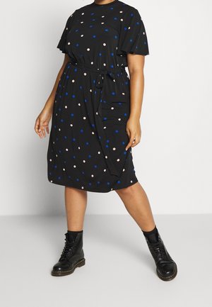 Robe noire jusqu'aux genoux avec des pois bleus et beiges, manches courtes, ceinture nouée à la taille, portée avec des bottines noires à lacets.