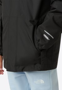 Veste imperméable noire avec une finition lisse, des poignets élastiques et un logo réfléchissant sur la manche. Associée à un pantalon bleu clair.