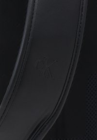 Cinturino in pelle nera con logo 'cK' impresso, caratterizzato da una superficie liscia e dettagli di cucitura fine. Sullo sfondo è visibile un motivo a rete.