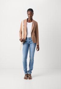 Blazer beige, top aderente bianco, jeans azzurri chiari e sandali con cinturini. Il blazer presenta un bottone singolo e una vestibilità leggermente sagomata.