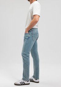 Jeans de denim azul claro con corte recto, que cuentan con cinco bolsillos y una cintura estándar, combinados con una camiseta blanca y zapatillas con rayas negras.