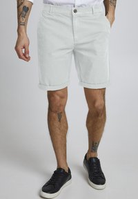 Pantalones cortos de algodón gris claro con dobladillos enrollados, cierre de botón y bolsillos laterales, combinados con zapatillas negras. Piel visible debajo de los pantalones cortos.