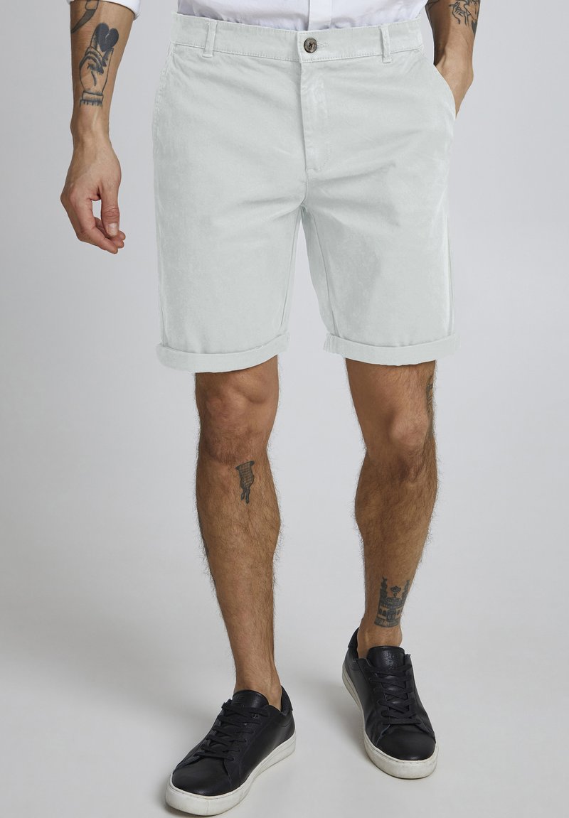 Pantalones cortos de algodón gris claro con dobladillos enrollados, cierre de botón y bolsillos laterales, combinados con zapatillas negras. Piel visible debajo de los pantalones cortos.
