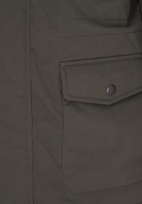 Primer plano de bolsillo con solapa de chaqueta de tela oscura, asegurado por un botón negro redondo con texto grabado y diseño de ancla.