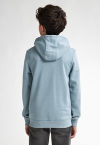 Ljusblå hoodie med snörning i huvan, långa ärmar, ribbade mudd och nederkant, tillverkad av mjukt material, bärs med mörka jeans.