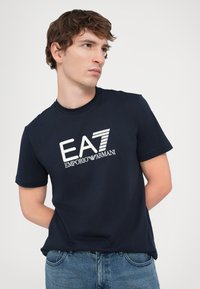 T-shirt in cotone blu navy con logo EA7 Emporio Armani bianco in un mix di testo in grassetto e a righe. Collo rotondo e maniche corte.