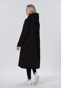 Cappotto lungo nero con cappuccio, caratterizzato da un design a strati e maniche lunghe. Abbinato a leggings neri e sneakers bianche. Texture liscia.