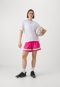 Hvit overdimensjonert t-skjorte med svart logo, rosa sportshorts med hvite og gullstriper, og svarte joggesko med hvite detaljer, holder en basketball.