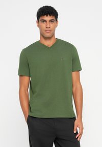 Ελαιοπράσινο μπλουζάκι V-neck από βαμβάκι, με κοντά μανίκια και μικρό λογότυπο στο στήθος, συνδυασμένο με μαύρο παντελόνι.