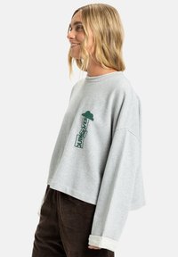 Sweatshirt gris au design court, avec un imprimé graphique vert. Il a un col rond et des poignets blancs sur les manches.