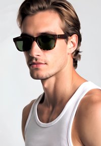 Occhiali da sole con montatura in tartaruga marrone e lenti verde scuro. Design rettangolare slim, con accenti sottili sulle tempie.