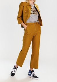 Veste jaune moutarde avec deux poches poitrine, t-shirt graphique gris, pantalons jaune moutarde, et baskets noires/bleues avec des lacets blancs.