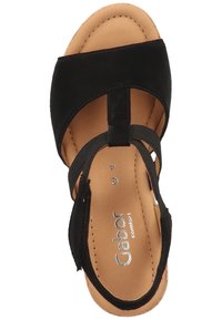 Sandale en daim noir avec un design à lanières, bride de cheville réglable et semelle intérieure rembourrée sur une base beige. Détail du logo métallique inclus.