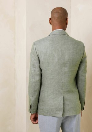 Mann trägt hellgrünen strukturierten Blazer und hellgraue Hose, steht mit dem Gesicht zu einer beigen Wand, die Hände entspannt an den Seiten.
