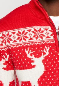 Czerwony sweter robiony na drutach z zamkiem w dekolcie, z białymi wzorami reniferów i płatków śniegu na klatce piersiowej, o teksturowanej powierzchni.