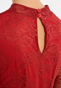 Haut en dentelle rouge avec un col montant et une ouverture en forme de clé dans le dos, présentant deux boutons rouges. Motifs floraux et design texturé visibles.