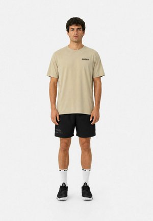 Mand stående iført beige kortærmet skjorte med teksten "CUERA", sorte shorts, hvide sokker med sort tekst og sorte sportssko.