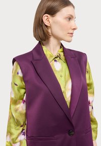 Lilla skreddersydd vest med en skarp krage og enkel knappelukking, parret med en grønn blomstermønstret bluse. Jevn stofftekstur.