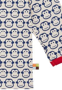 Tessuto in cotone con volti di scimmia blu ripetuti su uno sfondo crema, con accenti rossi ai bordi delle maniche e un'etichetta colorata.