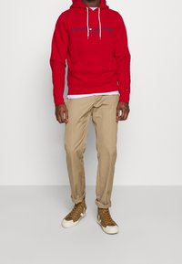 Homme portant un hoodie rouge Tommy Hilfiger, un pantalon beige retroussé aux chevilles et des baskets montantes marron avec des semelles blanches, se tenant contre un fond blanc.