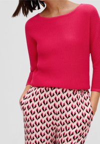 Fuchsia Rippstrickpullover mit weitem Ausschnitt, kombiniert mit gemusterten Hosen, die ein pinkes und schwarzes Pfeilmuster auf hellem Hintergrund zeigen.