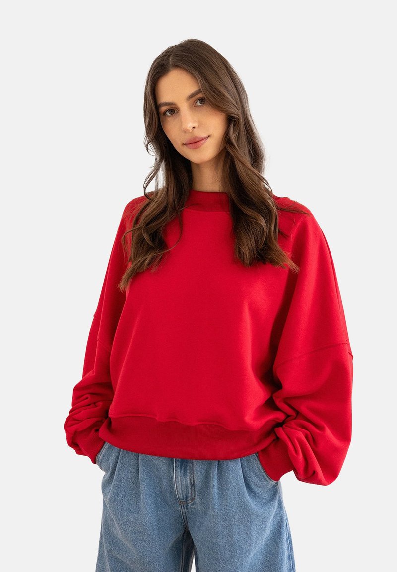 Czerwony sweter z opadającymi ramionami, prążkowanymi mankietami i luźnym krojem. Połączony z jasnoniebieskimi dżinsami, uwydatniającymi miękką teksturę materiału.