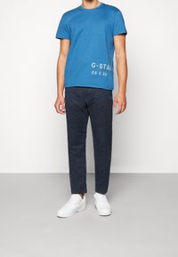 Blå bomullströja med vitt tryckt logotyp, kombinerad med mörkgrå chinos och vita sneakers. Avslappnad passform, rund halsringning, korta ärmar.