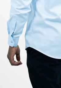 Lichtblauw overhemd met lange mouwen, voorzien van knoopmanchetten en een gebogen zoom, gecombineerd met donkere broek. Soepele stofstructuur.