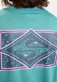 Tyrkysové tričko s krátkým rukávem, které na zadní straně obsahuje velkou grafiku složenou z diamantového tvaru s prasklým povrchem, obrysem v růžové barvě.