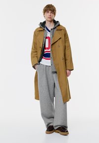 Bež plašč dvojnim zapenjanjem preko sivih oversized hlač. Plast se dopolnjuje s karirasto srajco in grafičnim puloverjem. Rjave čevlje dopolnjujejo videz.