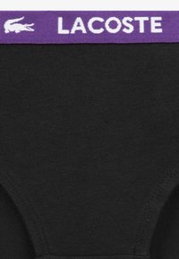Juodi bokseriai su lygiu paviršiumi ir violetine elastine juosta juosmens srityje, ant kurios baltomis raidėmis parašytas prekinis ženklas „LACOSTE“ ir logotipas.