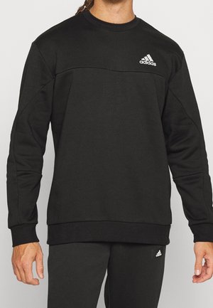 Mężczyzna w czarnej bluzie Adidas i dopasowanych czarnych spodniach, oba z białym logo Adidas na piersi i udzie.