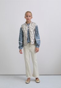 Chaleco acolchado plateado sobre una camisa azul de manga larga con estampado floral, combinado con vaqueros blancos acampanados y bailarinas doradas. Diseño sencillo y moderno.