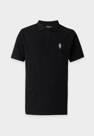 Homme debout portant un polo noir, un pantalon gris ample et des chaussures noires sur un fond clair uni.