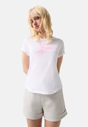 Jeune femme aux cheveux blonds portant un t-shirt blanc avec un motif rose et un short beige clair retroussé, debout devant un fond uni.