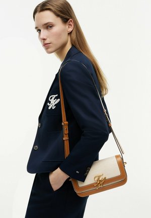 Młoda kobieta w granatowej marynarce z białym logo na kieszeni niesie beżowo-brązową skórzaną torbę na ramię ze złotym detalem logo.