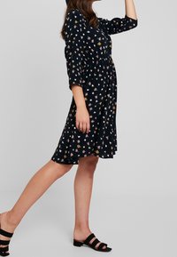 Blumendruck, knielanges Kleid aus navy Stoff mit dreiviertelÄrmeln. Hat eine geraffte Taille und einen lockeren Rock. Getragen mit schwarzen Sandalen.