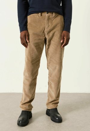 Mann trägt beige Cordhose, schwarzes Langarmshirt und schwarze Stiefel, steht auf grauem Boden vor einer schlichten hellen Wand.
